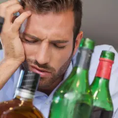 Mann mit Kopfschmerzen und leeren Flaschen. Was hilft gegen Kater? Viel Wasser trinken und Ruhe.