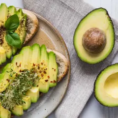 Avocado gesund: Toast mit frischen Avocadoscheiben, Sprossen und Gewürzen, daneben eine aufgeschnittene Avocado.
