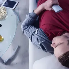 Mann in Midlife-Crisis liegt erschöpft auf dem Sofa, umgeben von Popcorn, Bier und einer Spielekonsole.