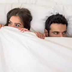 Ein Paar versteckt sich unter der Decke. Sie schauen sich an, als ob sie ein Geheimnis teilen oder darauf warten, dass etwas passiert. Wer wird wohl schneller kommen als Mann?