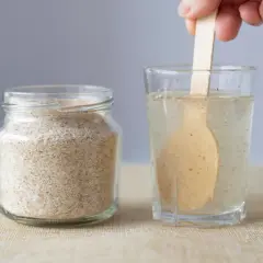 Glas mit Flohsamenschalen neben einem Glas Wasser, in dem ein Holzlöffel rührt. Richtige flohsamenschalen dosierung bei verstopfung ist wichtig.