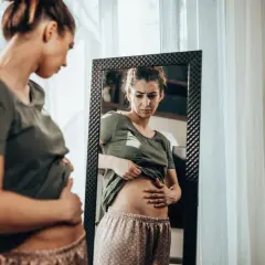 Frau mit besorgtem Gesichtsausdruck prüft ihren Bauch im Spiegel. Sie fragt sich: Luft im Bauch, was tun?