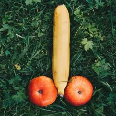 Eine Banane und zwei Äpfel liegen auf grünem Gras und bilden die Form eines kurzen dicken Penis.