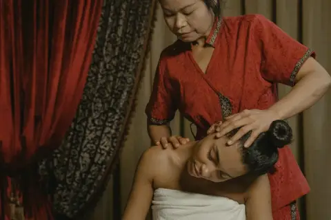 Eine Frau in einem roten Gewand genießt eine entspannende Thai-Massage. Ihre Augen sind geschlossen, während die Masseurin sanft ihren Kopf und Nacken behandelt.