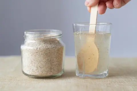 Glas mit Flohsamenschalen neben einem Glas Wasser, in dem ein Holzlöffel rührt. Richtige flohsamenschalen dosierung bei verstopfung ist wichtig.
