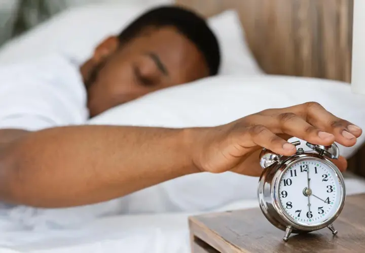 Ein Mann schaltet den Wecker aus. Warum kriegt man eine Morgenlatte? Vielleicht wegen des erholsamen Schlafs, der hier angedeutet wird.