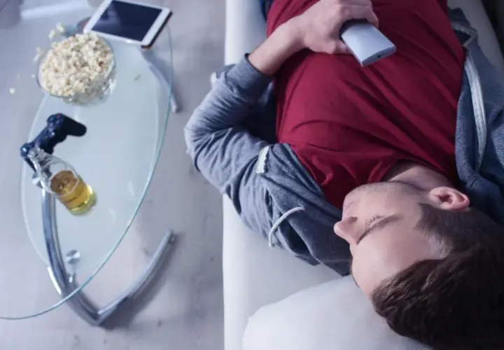 Mann in Midlife-Crisis liegt erschöpft auf dem Sofa, umgeben von Popcorn, Bier und einer Spielekonsole.