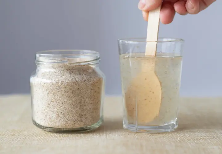 Glas mit Flohsamenschalen neben einem Glas Wasser, in dem ein Holzlöffel rührt. Richtige flohsamenschalen dosierung bei verstopfung ist wichtig.