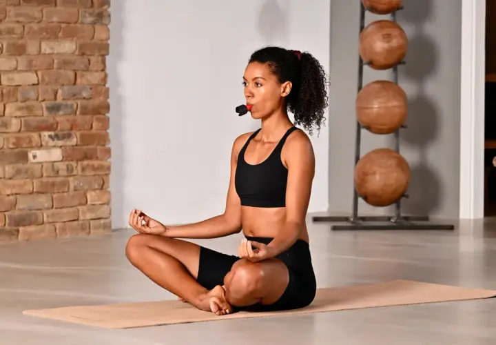 Frau in Schwarz übt neurozentriertes Training mit Atemtrainer. Sie sitzt im Lotussitz auf einer Matte.