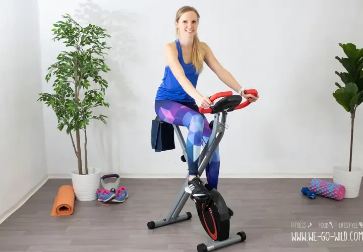Frau auf Heimtrainer Fahrrad, positive Erfahrungen beim Abnehmen. Trainingsmatte, Gewichte und Schuhe liegen bereit.