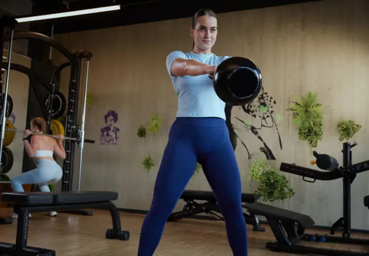 Frau macht Kettlebell-Schwünge als Teil ihres funktionellen Trainings. Im Hintergrund macht eine andere Frau Kniebeugen.