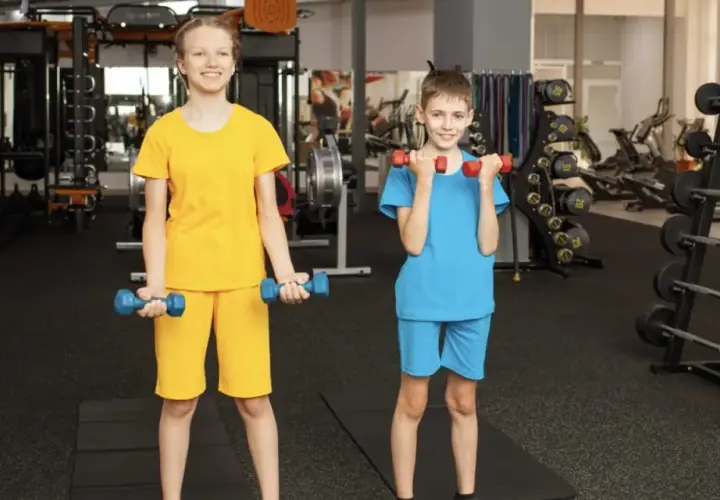 Zwei Kinder trainieren im Fitnessstudio mit Hanteln. Ab wie vielen Jahren darf man ins Gym? Hier wird gezeigt, dass auch junge Leute fit werden können!