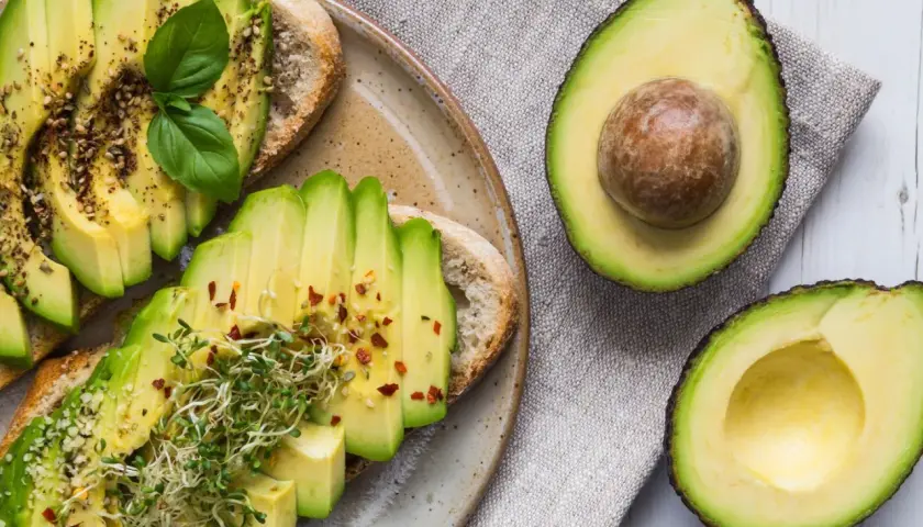 Avocado gesund: Toast mit frischen Avocadoscheiben, Sprossen und Gewürzen, daneben eine aufgeschnittene Avocado.