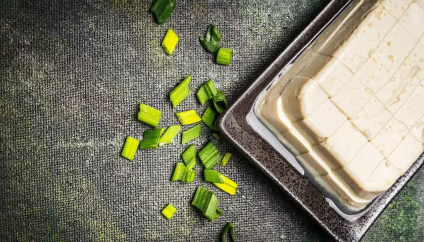 Seidentofu, zart und cremig, liegt auf einem dunklen Teller, umgeben von frischen Frühlingszwiebeln. Perfekt für leichte Gerichte.