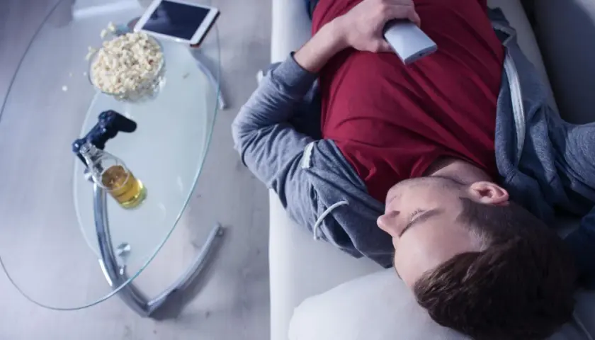 Mann in Midlife-Crisis liegt erschöpft auf dem Sofa, umgeben von Popcorn, Bier und einer Spielekonsole.