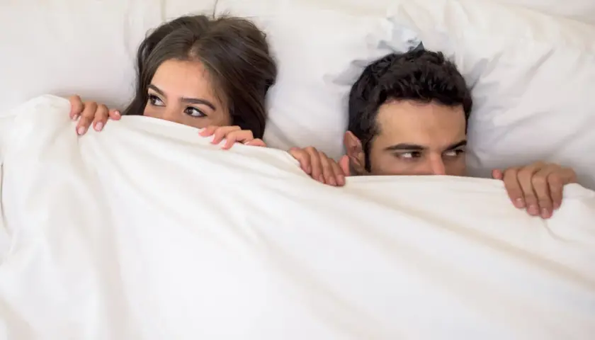 Ein Paar versteckt sich unter der Decke. Sie schauen sich an, als ob sie ein Geheimnis teilen oder darauf warten, dass etwas passiert. Wer wird wohl schneller kommen als Mann?