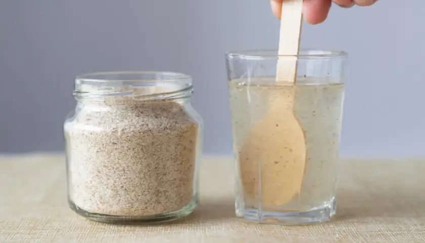 Glas mit Flohsamenschalen neben einem Glas Wasser, in dem ein Holzlöffel rührt. Richtige flohsamenschalen dosierung bei verstopfung ist wichtig.