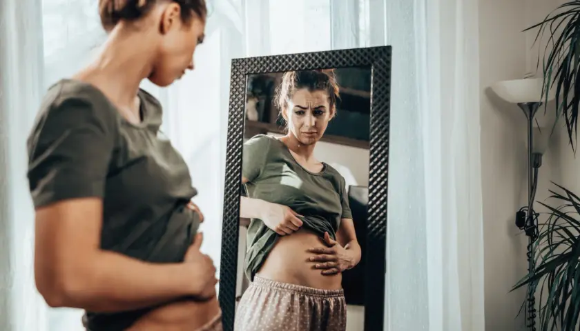 Frau mit besorgtem Gesichtsausdruck prüft ihren Bauch im Spiegel. Sie fragt sich: Luft im Bauch, was tun?