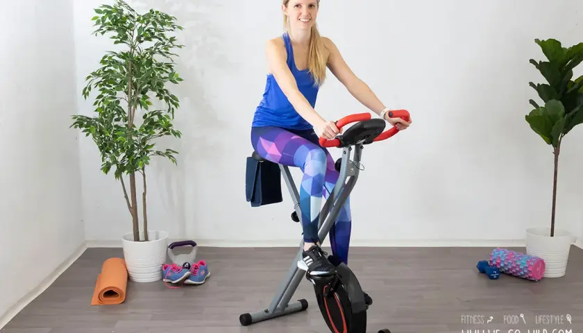 Frau auf Heimtrainer Fahrrad, positive Erfahrungen beim Abnehmen. Trainingsmatte, Gewichte und Schuhe liegen bereit.