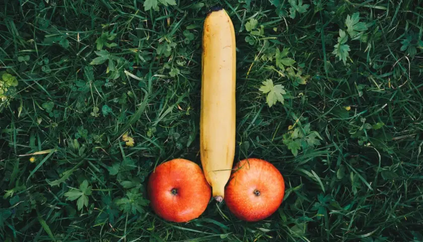 Eine Banane und zwei Äpfel liegen auf grünem Gras und bilden die Form eines kurzen dicken Penis.