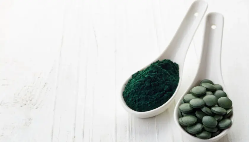 Zwei Löffel mit Spirulinapulver und Tabletten. Die Spirulina Wirkung ist vielfältig, von Energie bis zur Entgiftung.