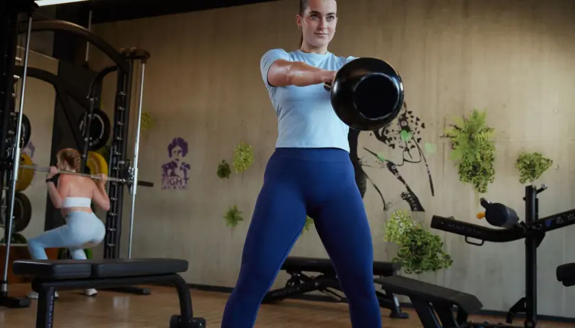 Frau macht Kettlebell-Schwünge als Teil ihres funktionellen Trainings. Im Hintergrund macht eine andere Frau Kniebeugen.