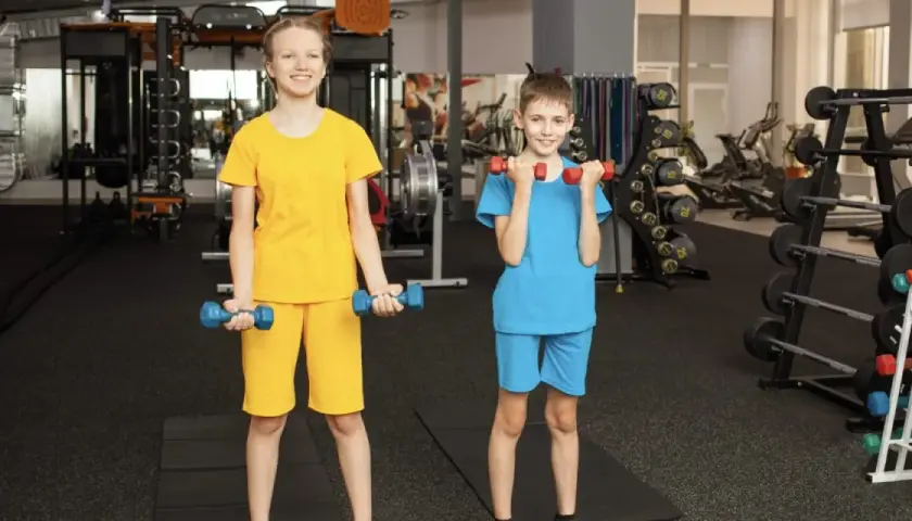 Zwei Kinder trainieren im Fitnessstudio mit Hanteln. Ab wie vielen Jahren darf man ins Gym? Hier wird gezeigt, dass auch junge Leute fit werden können!