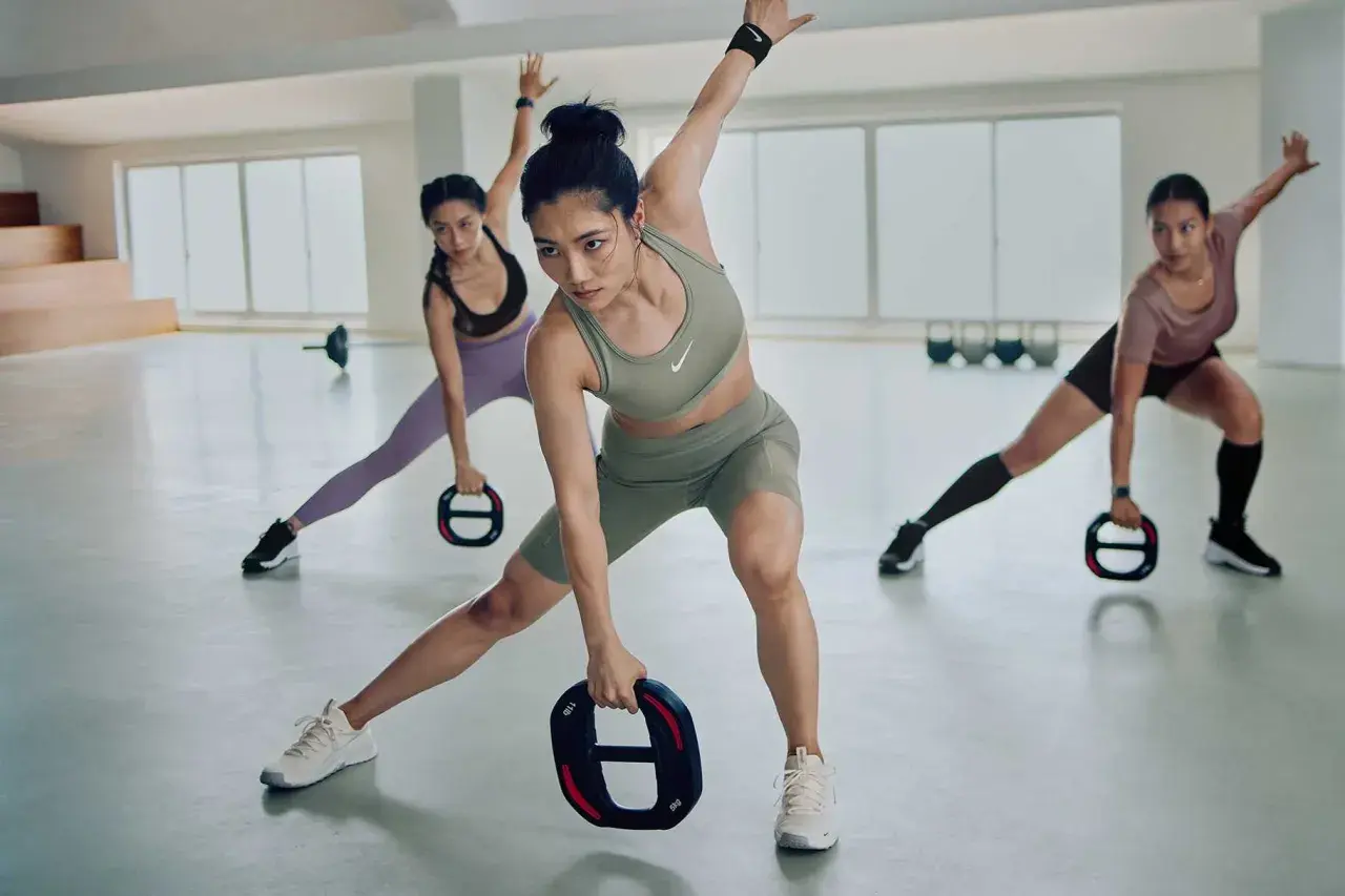 Drei Frauen in Sportkleidung trainieren in einer hellen Halle. Sie halten Gewichte und machen Ausfallschritte, um ihre aerobe Zone zu aktivieren.