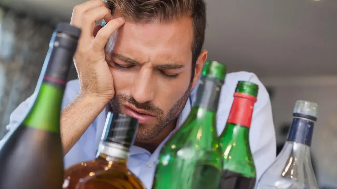 Mann mit Kopfschmerzen und leeren Flaschen. Was hilft gegen Kater? Viel Wasser trinken und Ruhe.
