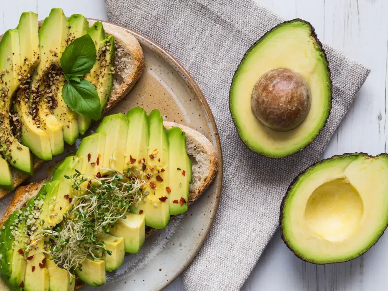Avocado gesund: Toast mit frischen Avocadoscheiben, Sprossen und Gewürzen, daneben eine aufgeschnittene Avocado.