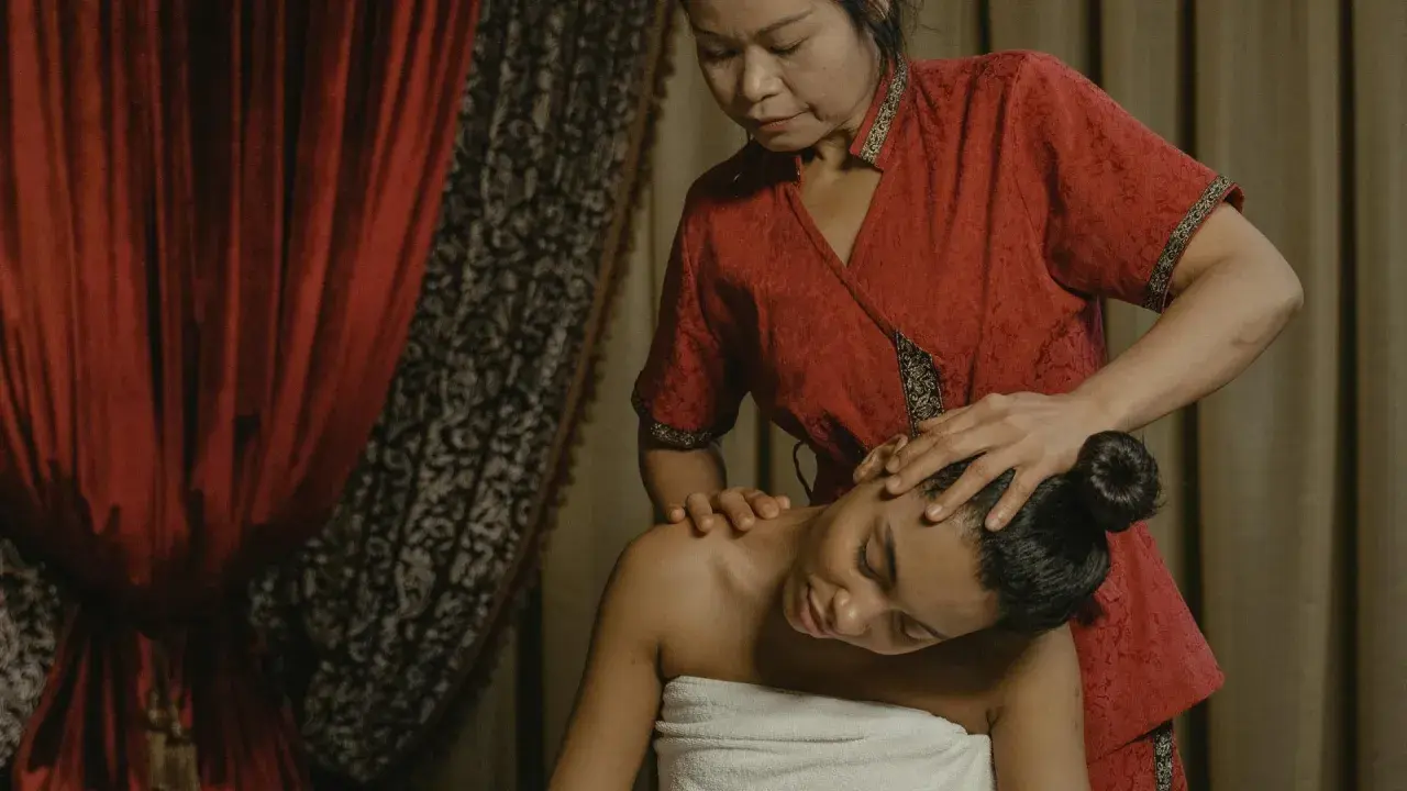 Eine Frau in einem roten Gewand genießt eine entspannende Thai-Massage. Ihre Augen sind geschlossen, während die Masseurin sanft ihren Kopf und Nacken behandelt.