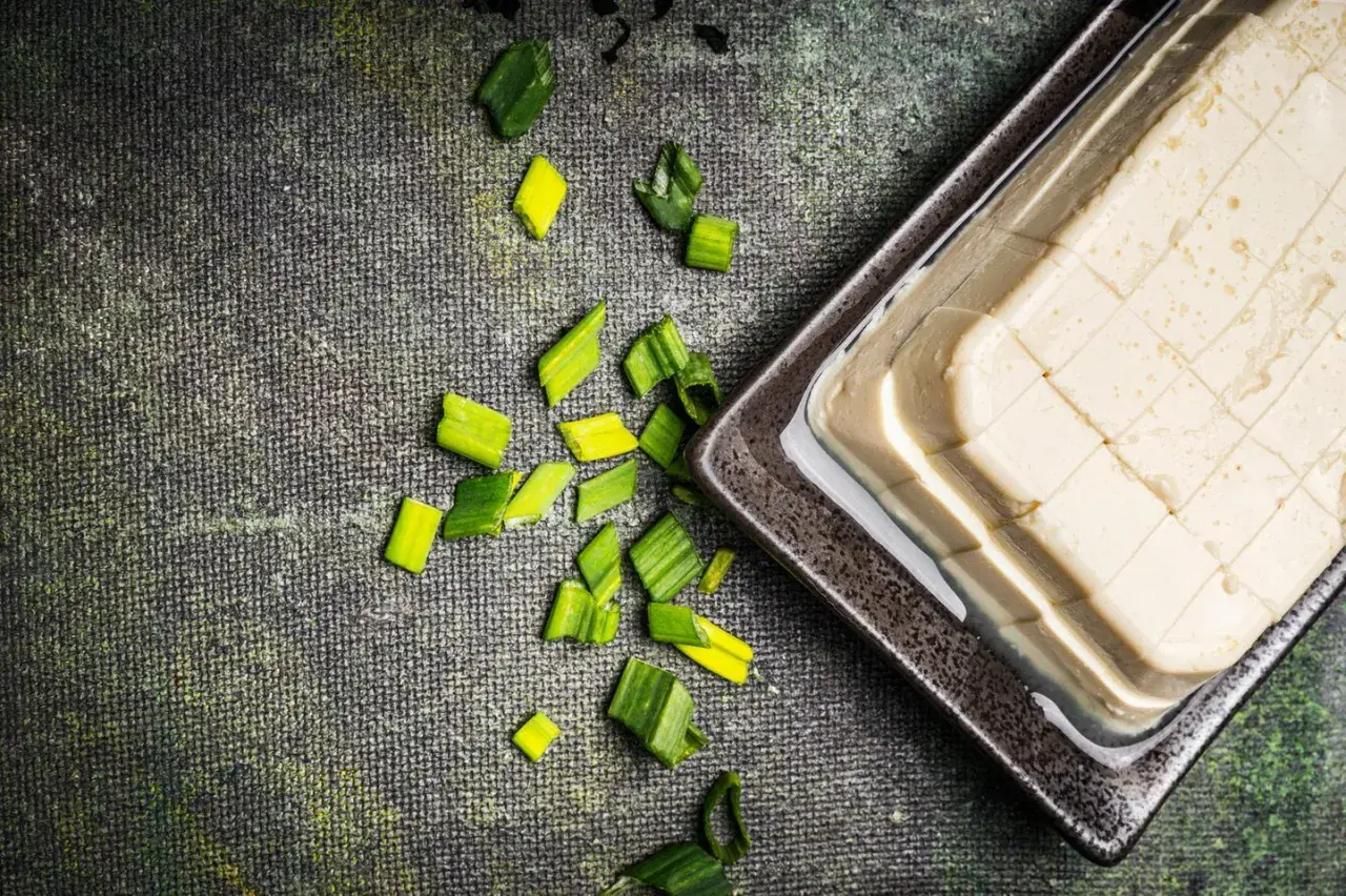 Seidentofu, zart und cremig, liegt auf einem dunklen Teller, umgeben von frischen Frühlingszwiebeln. Perfekt für leichte Gerichte.