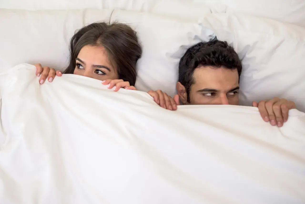 Ein Paar versteckt sich unter der Decke. Sie schauen sich an, als ob sie ein Geheimnis teilen oder darauf warten, dass etwas passiert. Wer wird wohl schneller kommen als Mann?