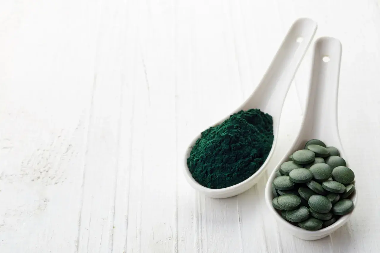 Zwei Löffel mit Spirulinapulver und Tabletten. Die Spirulina Wirkung ist vielfältig, von Energie bis zur Entgiftung.