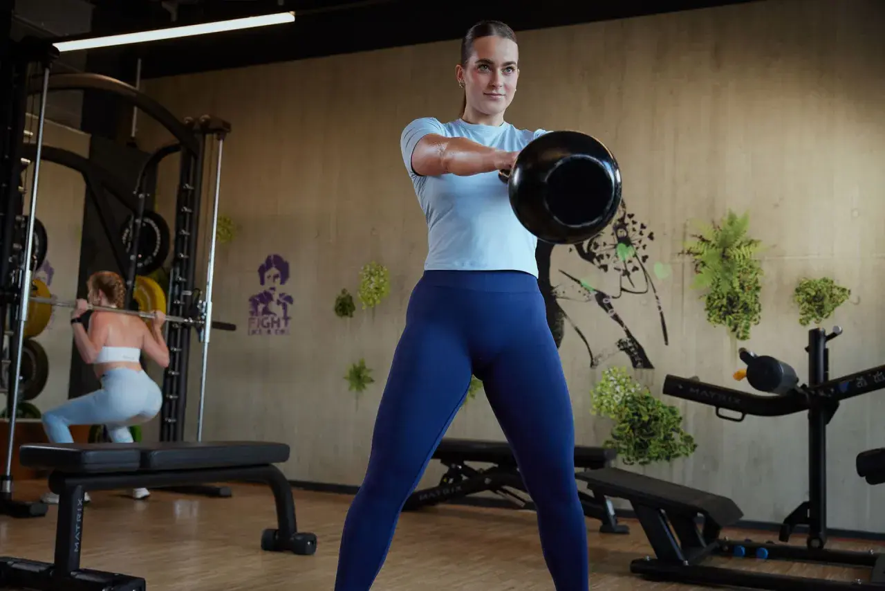 Frau macht Kettlebell-Schwünge als Teil ihres funktionellen Trainings. Im Hintergrund macht eine andere Frau Kniebeugen.