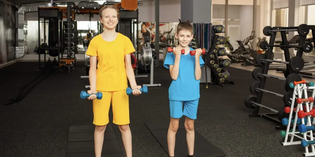 Zwei Kinder trainieren im Fitnessstudio mit Hanteln. Ab wie vielen Jahren darf man ins Gym? Hier wird gezeigt, dass auch junge Leute fit werden können!