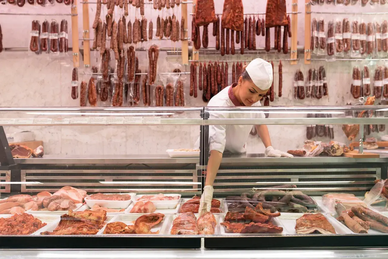 Fleischtheke mit Wurst und Fleischwaren. Eine Verkäuferin sortiert Ware. Hier findet man Inspiration, wie viel Fleisch pro Woche auf dem Teller landen könnte.