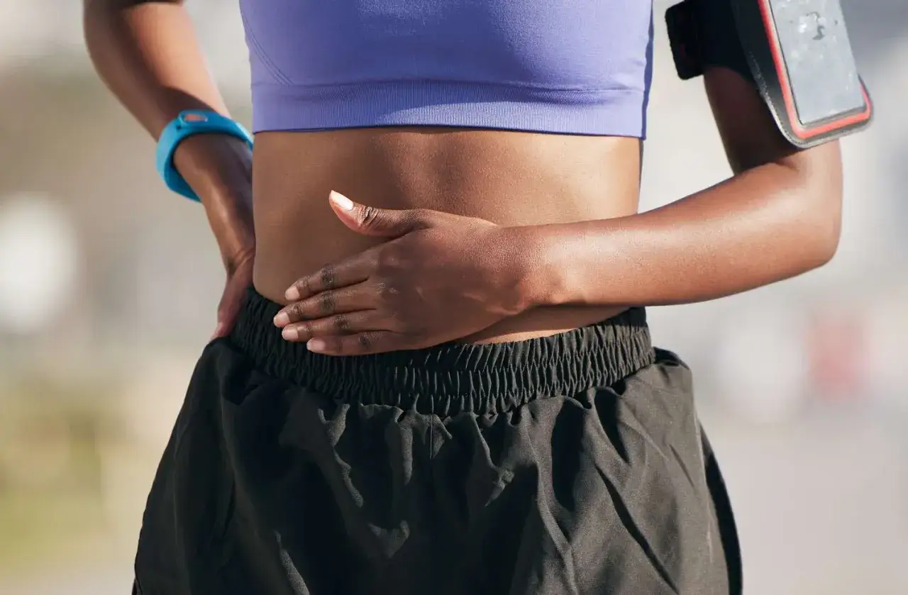 Läuferin hält sich den Bauch, vielleicht wegen zu viel Kreatin. Sie trägt Sportkleidung und eine Armbinde für ihr Handy.