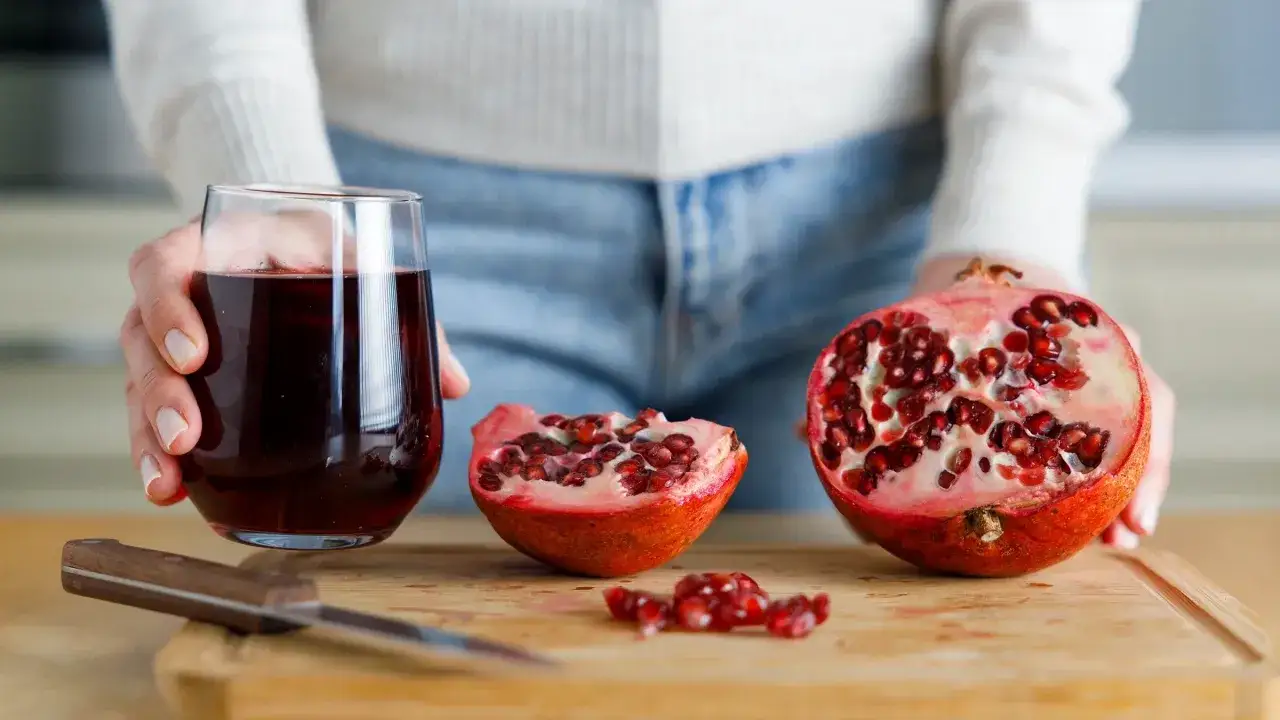 Eine Hand h&auml;lt ein Glas Granatapfelsaft. Daneben liegen auf einem Holzbrett ein aufgeschnittener Granatapfel und Kerne. Granatapfel gesund!