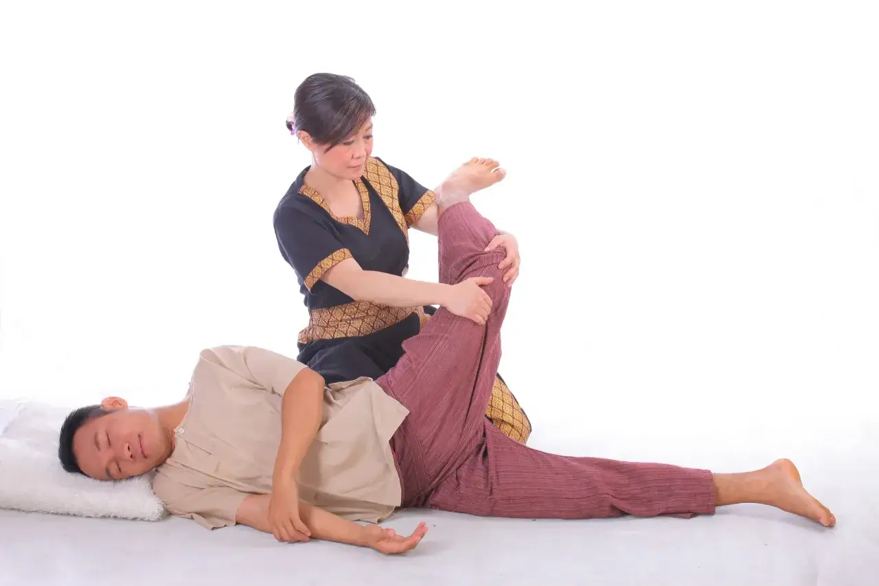 Eine Frau in traditioneller Kleidung führt eine Thai-Massage durch, indem sie das Bein eines liegenden Mannes sanft streckt.