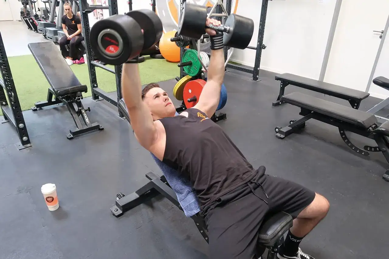 Mann hebt schwere Hanteln bei einem incline bench press. Ein Getränkebecher steht auf dem Boden.