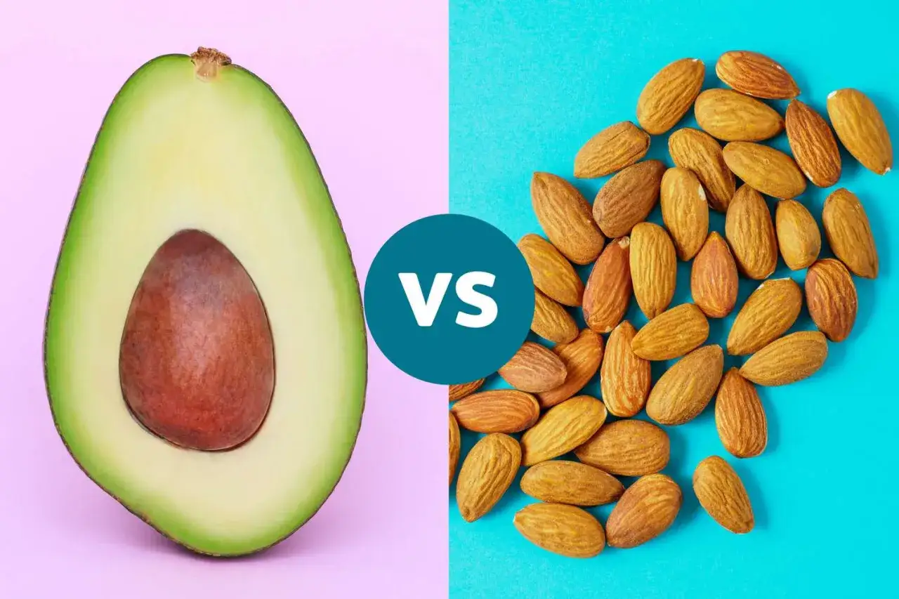 Halbe Avocado mit Kern auf rosa Hintergrund, daneben Mandeln auf blauem Hintergrund. Avocado gesund vs. Mandeln.