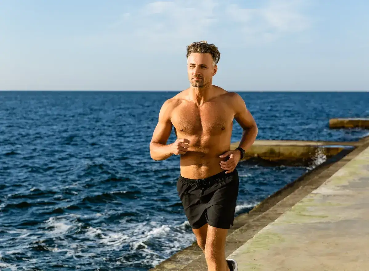 Ein normal trainierter Mann läuft am Meer entlang. Die Sonne scheint, das Meer ist blau.