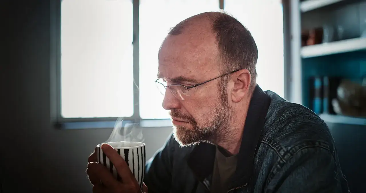 Ein nachdenklicher Mann mit Brille h&auml;lt eine dampfende Tasse. Er wirkt wie ein Mann in der Midlife-Crisis, der &uuml;ber das Leben sinniert.