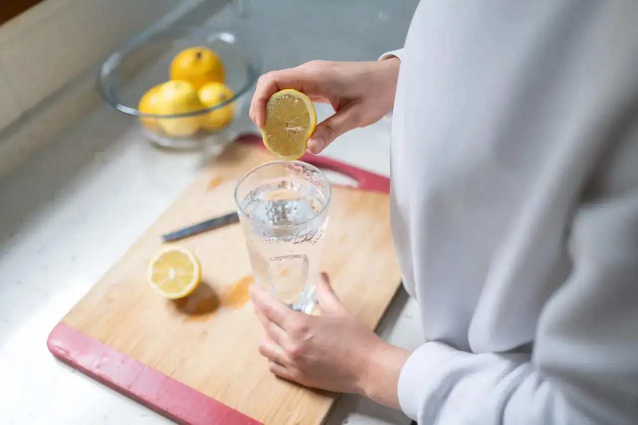 Eine Hand presst eine Zitronenhälfte über ein Glas Wasser. Frisches zitronenwasser am morgen für einen guten Start.