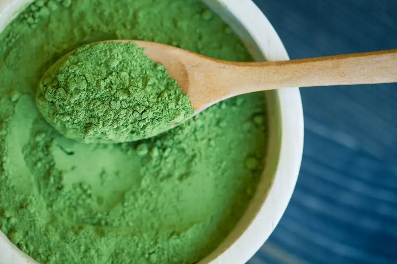 Holzschale mit feinem, gr&uuml;nem Spirulinapulver. Ein Holzl&ouml;ffel hebt eine Portion hervor, die die positive Spirulina Wirkung andeutet.