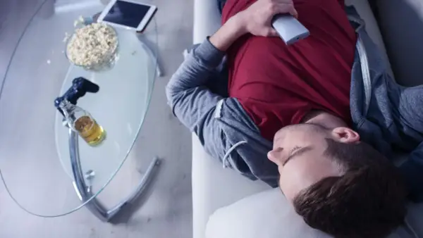 Mann in Midlife-Crisis liegt erschöpft auf dem Sofa, umgeben von Popcorn, Bier und einer Spielekonsole.